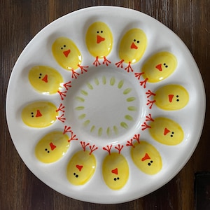 May include: A white ceramic egg plate with yellow chick-shaped egg holders. Each chick has a red beak and feet, and black eyes. The center of the plate has a ring of green painted leaves.