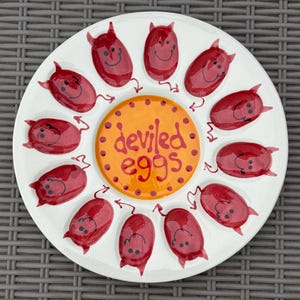 May include: White ceramic deviled egg serving platter with a central orange circle and the words "deviled eggs." The platter features red devil-shaped egg holders around the perimeter, each with a smiling face and horns.