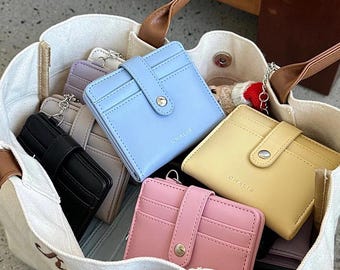 Puede incluir: Una bolsa de lona llena de varias carteras pequeñas. Las carteras son de diferentes colores, incluyendo azul claro, amarillo, rosa y gris. Cada cartera tiene un cierre a presión y la palabra "CHAKIB" grabada en la parte delantera.