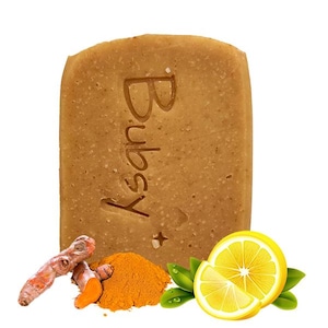 Puede incluir: Una pastilla de jabón rectangular de color marrón claro con la palabra "Bubsy" grabada. Junto al jabón hay trozos de raíz de cúrcuma, un montón de polvo de cúrcuma y un limón en rodajas con hojas verdes.