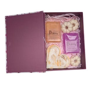 Puede incluir: Una caja de regalo con exterior morado e interior floral, que contiene jabones y objetos decorativos. La caja contiene dos jabones rectangulares, dos jabones blancos con forma de calabaza y un jabón en forma de corazón. Los jabones están etiquetados con la marca "Bubsy".