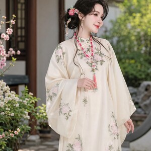 May include: A cream-colored long-sleeved dress featuring delicate floral embroidery in shades of pink and green. The garment has a high collar, wide sleeves, and a flowing silhouette, accented by a long beaded necklace with a pink tassel pendant.