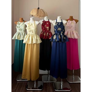 May include: A selection of halter-neck dresses in shades of teal, yellow, red, navy, and pink. Each dress features a peplum top with floral details and a long, flowing skirt. The dresses are displayed on mannequins, showcasing their design.