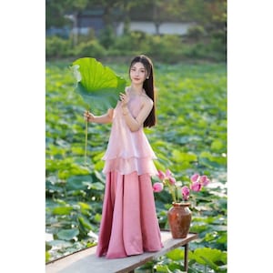 May include: A pink halter-neck dress with layered ruffles and a long skirt. The dress is worn outdoors, with a large green leaf held in the hand. A vase of pink flowers sits nearby. The dress is made of a soft, flowing fabric.