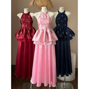 May include: Three halter-neck dresses in burgundy, pink, and navy blue. Each dress features a high collar, decorative buttons, and a layered peplum. The burgundy and navy dresses have a subtle pattern. The dresses are displayed on mannequins.