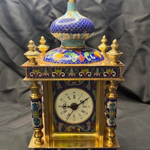 May include: An ornate, gold-toned clock with a domed top and intricate enamel detailing. The clock face is white with black Roman numerals and hands. The clock's design features blue, green, and red floral patterns, and it stands approximately 10 inches tall.