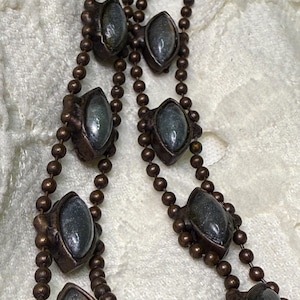 Puede incluir: Primer plano de un collar vintage con dos hilos de pequeñas cuentas redondas de color marrón oscuro. Cada hilo tiene piedras ovaladas de color gris oscuro engastadas en marcos de metal ornamentados de color bronce. El collar se muestra sobre una superficie blanca texturizada.