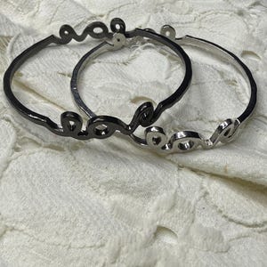 May include: Two metal bangle bracelets, one in a polished silver tone and one in a dark gunmetal finish. Each bracelet features the word love written in a continuous cursive script that forms the circular band of the jewelry piece.