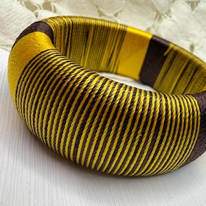 Puede incluir: Un brazalete grueso y redondo con un diseño llamativo. La pulsera está envuelta en bandas alternas de hilos amarillos y marrón oscuro. Las bandas amarillas son más anchas, creando un patrón audaz y llamativo. Una pieza de declaración.