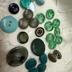 May include: A collection of various buttons in shades of green, blue, and brown. The buttons vary in size, shape, and material, including round, oval, and fabric-covered options. Some buttons have two or four holes, while others have a shank.