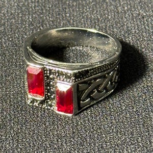 Könnte beinhalten: Silberfarbener Ring mit zwei rechteckigen, leuchtend roten Edelsteinen. Der Ring hat ein aufwendiges Design mit einem strukturierten Band und dekorativen Mustern. Ein stilvolles Schmuckstück.