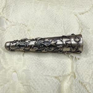 Könnte beinhalten: Ein silberfarbener, zylindrischer Flaschenverschluss mit detailliertem Blumen- und Blattdesign. Der Verschluss hat eine konische Form und ist mit aufwendigen Schnitzereien von Weinreben, Blättern und kleinen Beeren verziert. Das Metall hat ein leicht gealtertes Aussehen.