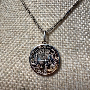 May include: Silver-toned pendant necklace featuring a round charm with the text "3rd Millennium 2000". The charm depicts iconic landmarks and a rising sun design. The necklace has a delicate chain.