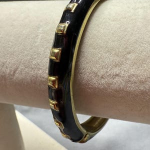 Puede incluir: Una pulsera de brazalete negra y dorada. La pulsera tiene un acabado de esmalte negro brillante con detalles cuadrados dorados. Los bordes de la pulsera también son dorados. La pulsera se muestra sobre una superficie blanca.