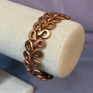 Puede incluir: Una pulsera de color cobre con un diseño repetitivo de bucles abiertos. La pulsera se muestra sobre una forma cilíndrica gris claro. Los bucles están conectados para formar una banda flexible. El metal tiene un tono cálido, rojizo-marrón.