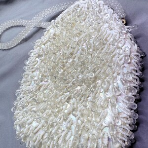 May include: A vintage white beaded handbag with a clear beaded handle. The bag is covered in small white beads and larger, clear, teardrop-shaped beads. The clasp is gold-toned.