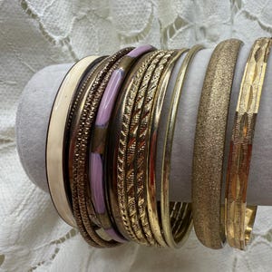 Puede incluir: Una colección de pulseras de oro y metal mixto. Las pulseras incluyen una variedad de texturas y diseños, incluyendo superficies lisas, texturizadas y estampadas. Algunas pulseras tienen un acabado esmaltado rosa claro o crema. Las pulseras se exhiben sobre una forma cilíndrica gris.