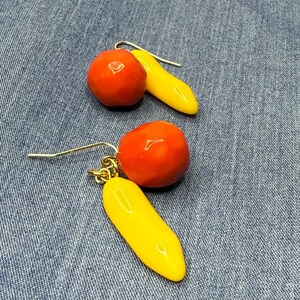 May include: Earrings with a red, spherical bead and a yellow, banana-shaped charm. The earrings are suspended from gold-coloured hooks. The background is a denim fabric.
