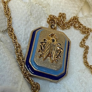 Op de afbeelding: Goudkleurige ketting met een rechthoekige hanger met een gouden insectenontwerp. De hanger heeft een blauwe en lichtblauwe emaille rand. De ketting heeft een delicate touwstijl. De ketting wordt gepresenteerd op een witte, getextureerde ondergrond.