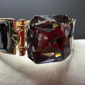 May include: A gold-toned hinged bracelet with a large, faceted, smoky-gray gemstone. The bracelet features a black enamel section and a gold clasp. The gemstone is rectangular with multiple facets, reflecting light.