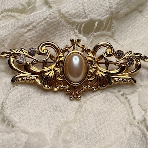 Op de afbeelding: Goudkleurige broche met een ovaal, parelachtig middenstuk en een sierlijk filigraanontwerp. De broche heeft kleine, heldere kristallen en een symmetrische, elegante vorm, geschikt voor formele gelegenheden of een vintage touch.