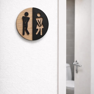 Könnte beinhalten: Rundes Toilettenschild aus Holz, geteilt in zwei Hälften. Eine Hälfte ist aus Naturholz mit einer männlichen Silhouette, die andere ist schwarz mit einer weiblichen Silhouette. Das Schild befindet sich an einer weißen, strukturierten Wand.