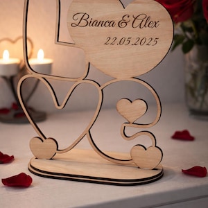 Pode incluir: Decoração de casamento em madeira com a palavra "LOVE" formada por corações. O coração superior é gravado com "Bianca & Alex" e a data "22.05.2025". A decoração está sobre uma superfície branca com pétalas de rosa vermelhas.