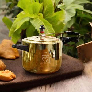 May include: A polished gold-tone pressure cooker with a reflective metallic finish, featuring black plastic side handles, a locking lid mechanism with a central pressure valve, and the text LEKA printed in white on the front of the cylindrical body.