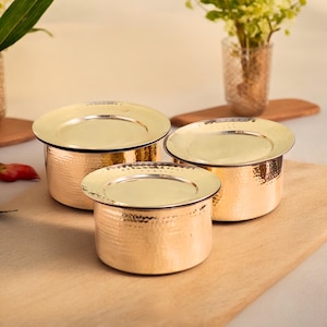 Puede incluir: Tres recipientes de metal martillado dorado con tapas. Los recipientes están anidados uno dentro del otro, con el recipiente más grande en la parte inferior y el más pequeño en la parte superior.