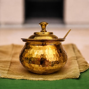 May include: A polished gold-toned metal jar with a hammered texture, featuring a rounded body, a matching lid with a central knob, and a small metal spoon resting inside. The container has a wide base and a narrow neck, reflecting light.