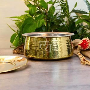 May include: A gold-colored, hammered-texture serving bowl with a silver rim. The bowl has an etched design on the front. A gold-colored plate is in the foreground. Green foliage is in the background.