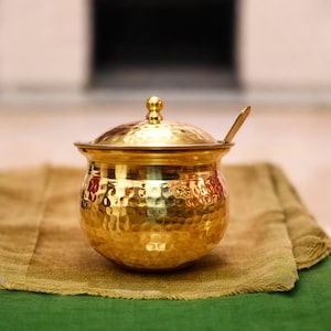 May include: A gold hammered metal container with a lid and a small spoon. The container is sitting on a brown fabric surface.