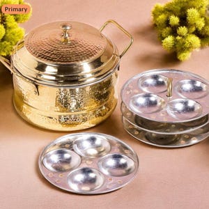 Könnte beinhalten: Ein goldfarbener Idli-Dampfgarer aus gehämmertem Metall mit Deckel und Griff, zusammen mit mehreren silberfarbenen Idli-Formen. Der Dampfgarer hat eine strukturierte Oberfläche und eine runde Form. Die Formen sind gestapelt.