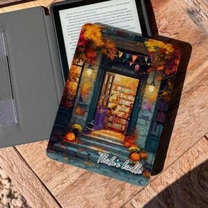 Könnte beinhalten: Ein Kindle E-Reader mit einer dekorativen Hülle, die eine Buchladen-Szene mit Kürbissen und Herbstlaub zeigt. Die Hülle ist geöffnet und zeigt den Bildschirm. Daneben liegt ein graues Etui.