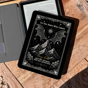 Può includere: Una custodia nera per Kindle con il testo "To The Stars Who Listen" e un disegno di montagna e stella. Sono visibili anche una custodia grigia per Kindle e un dispositivo Kindle con schermo illuminato. Su una superficie di legno.