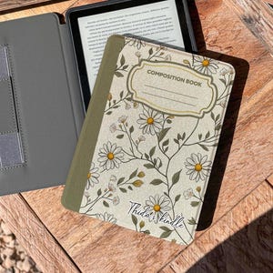 May include: A gray Kindle e-reader with a black screen, open to a page of text, next to a floral-patterned composition book cover. The cover features white daisies and green leaves on a cream background, with the text "COMPOSITION BOOK".