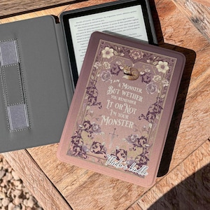 Op de afbeelding: Een Kindle e-reader met een decoratieve hoes met bloemen- en tekstelementen. De hoes heeft een citaat dat luidt: "Ik ben een monster, maar of je het je herinnert of niet, ik ben je monster". De e-reader zit in een grijze hoes.