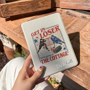 Peut inclure: Une housse de Kindle gris clair avec un motif sur le thème du hockey. La housse présente le texte "GET IN LOSER" et "We're going to THE COTTAGE", ainsi que des illustrations de joueurs de hockey et d'un palet. Le texte "Thida's kindle" est également visible.