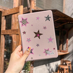 May include: A light pink e-reader case featuring a scattered pattern of various stars in different sizes and designs, including leopard print, solid pink, and black and white striped patterns. The name Thida is printed in a script font.