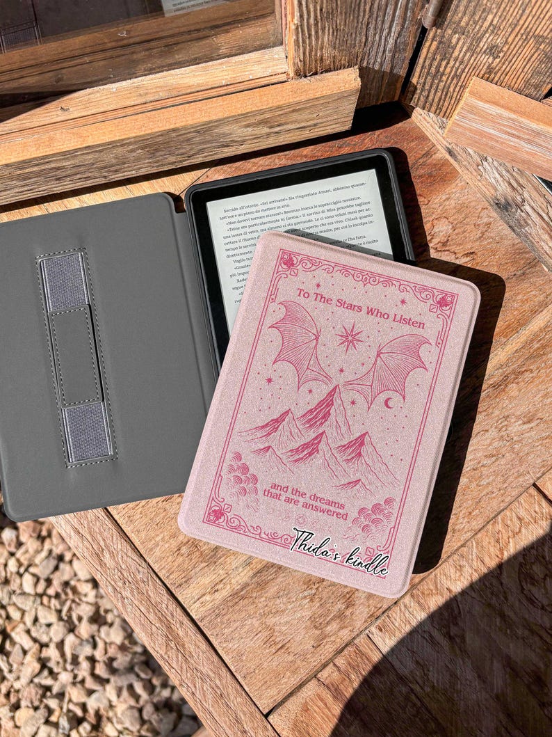 May include: A pink Kindle cover with the text "To The Stars Who Listen" and "and the dreams that are answered". The cover features a mountain and star design. A gray Kindle case is open next to it.