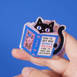Pode incluir: Um broche de acrílico transparente com um gato preto de olhos azuis grandes espreitando por cima de um livro azul aberto. A capa do livro apresenta padrões florais e o texto: HOW TO BUY NEW BOOKS & PRETEND LIKE IT WAS AN ACCIDENT.