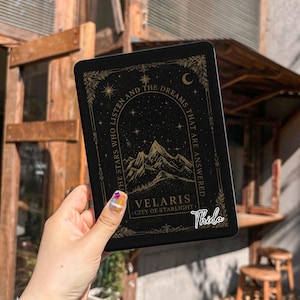 May include: A black e-reader skin featuring a gold-colored mountain range illustration, a starry night sky, and the text "THE STARS WHO LISTEN AND THE DREAMS THAT ARE ANSWERED" along with "VELARIS CITY OF STARLIGHT" and a cursive "Thida" signature.