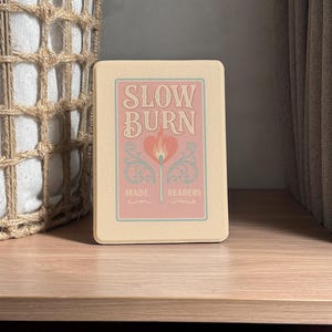 A retro-inspired Kindle case designed for book lovers, featuring the phrase "Slow Burn" in bold vintage lettering above a flaming heart-tipped match. Surrounded by elegant flourishes and the words "Made for Readers," the soft pink and cream palette adds a romantic, nostalgic charm. Includes a handy strap, magnetic back, and auto sleep/wake function—an ideal gift for readers who adore slow-burn romance stories and cozy book club nights.