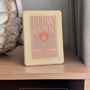 A charming Kindle case featuring vintage-style artwork of a matchbox labeled "Slow Burn" with a lit match topped with a heart. Below the flame, a ribbon banner reads "Made for Readers," celebrating fans of slow-burn romance. Set against a muted pink background, the case includes a convenient strap, magnetic back, and auto sleep/wake function—perfect for cozy, long reading sessions.