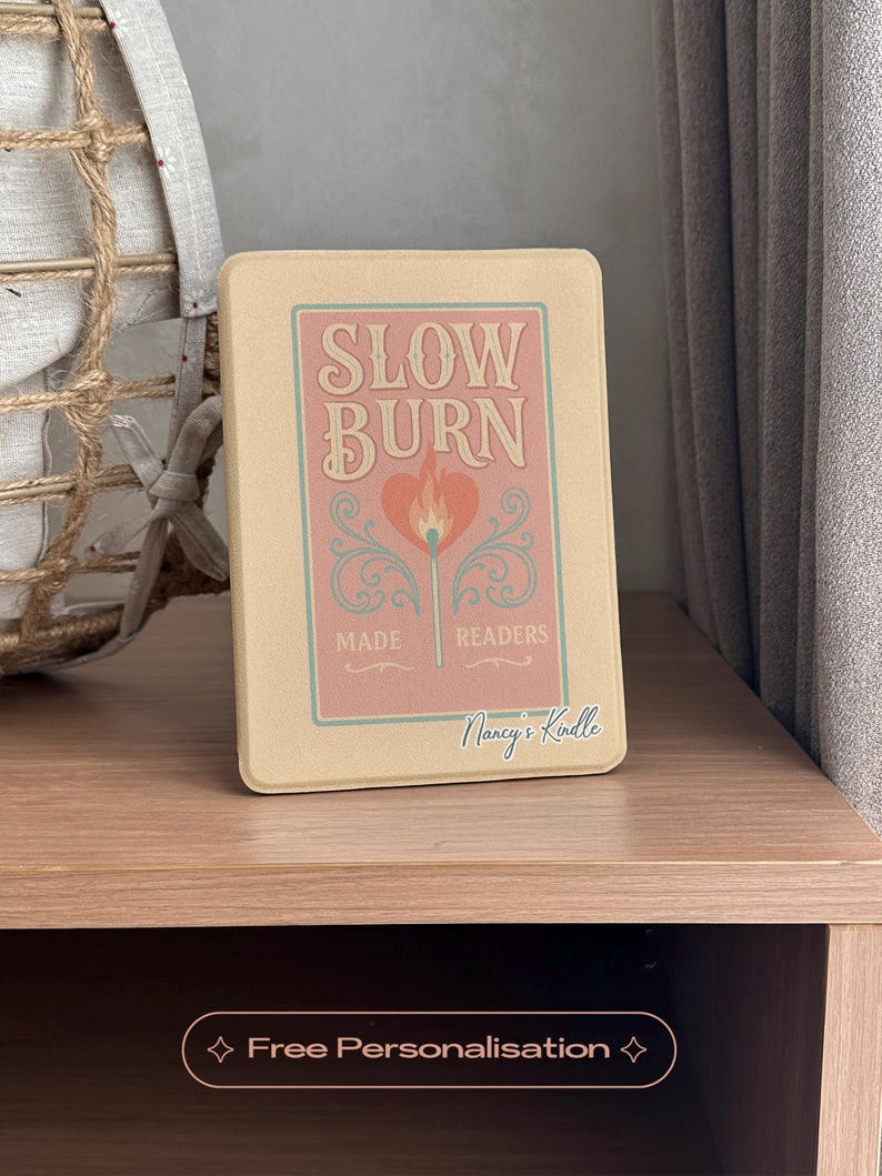 A charming Kindle case featuring vintage-style artwork of a matchbox labeled "Slow Burn" with a lit match topped with a heart. Below the flame, a ribbon banner reads "Made for Readers," celebrating fans of slow-burn romance. Set against a muted pink background, the case includes a convenient strap, magnetic back, and auto sleep/wake function—perfect for cozy, long reading sessions.