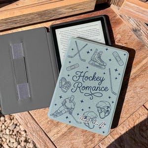 Peut inclure: Un étui gris pour Kindle avec une couverture bleu clair ornée d'illustrations sur le thème du hockey et du texte "Hockey Romance". L'étui est ouvert, révélant l'écran du Kindle. L'étui est posé sur une surface en bois.
