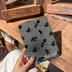 May include: A gray tablet case featuring a pattern of black dragon silhouettes and small white stars. The text "Theia's kindle" is printed in white script on the lower right corner. The case is held against a wooden background.