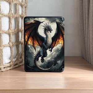 May include: A black case featuring a detailed illustration of a dragon with black and orange wings, soaring against a cloudy sky. The dragon has a blue body and a long tail. The case is displayed on a wooden surface.