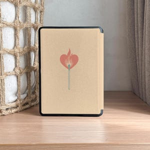 A charming Kindle case featuring vintage-style artwork of a matchbox labeled "Slow Burn" with a lit match topped with a heart. Below the flame, a ribbon banner reads "Made for Readers," celebrating fans of slow-burn romance. Set against a muted pink background, the case includes a convenient strap, magnetic back, and auto sleep/wake function—perfect for cozy, long reading sessions.back
