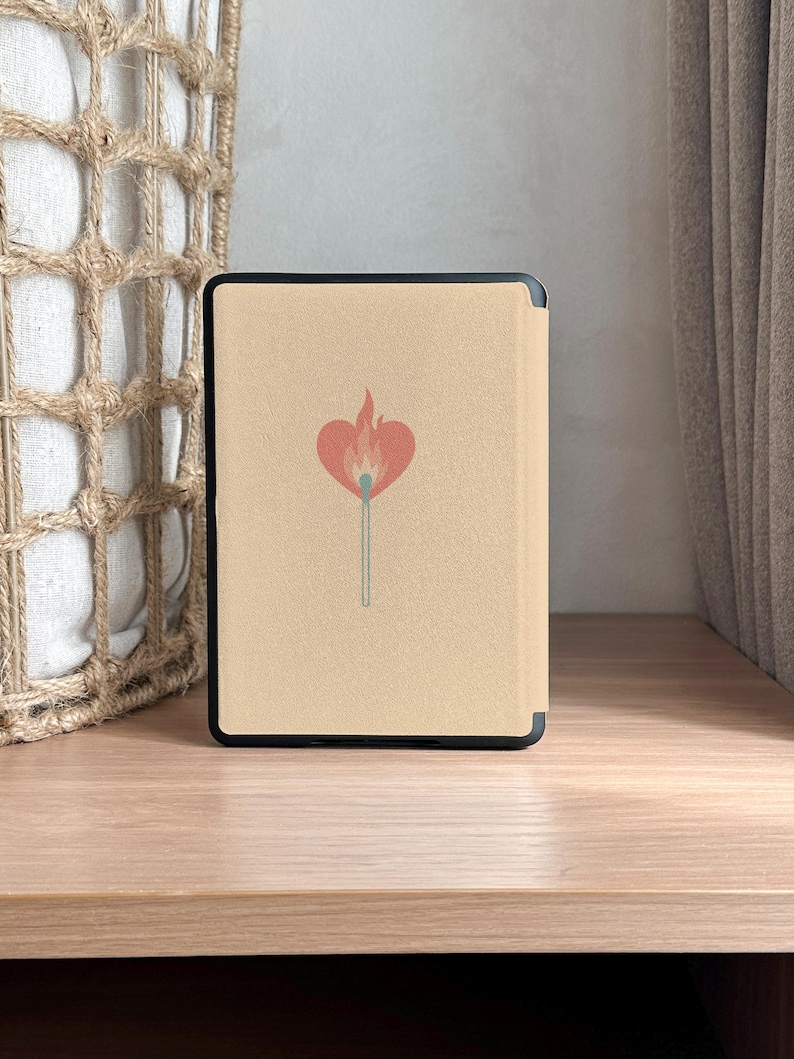 A charming Kindle case featuring vintage-style artwork of a matchbox labeled "Slow Burn" with a lit match topped with a heart. Below the flame, a ribbon banner reads "Made for Readers," celebrating fans of slow-burn romance. Set against a muted pink background, the case includes a convenient strap, magnetic back, and auto sleep/wake function—perfect for cozy, long reading sessions.back